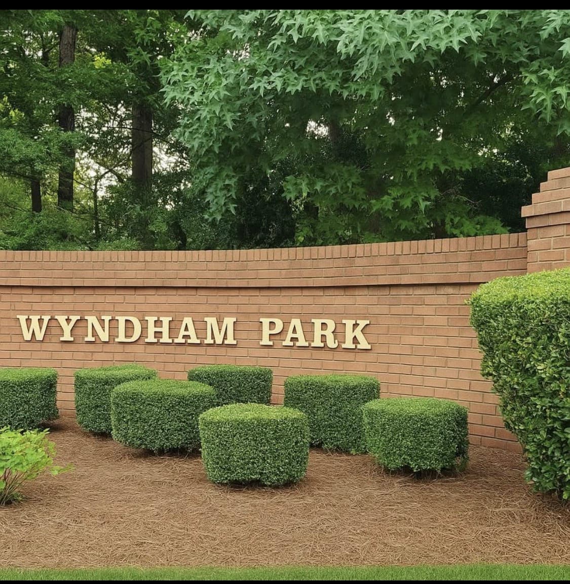 Wyndham Park entrance sign with brick wall, clipped shrubs, and mature trees